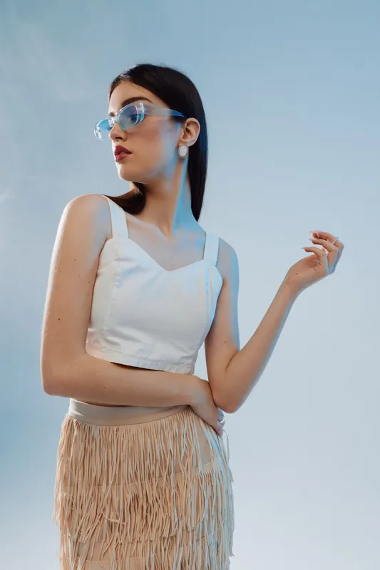 Young woman in white top and fringed skirt poses confidently wearing blue sunglasses against a clear sky. Concept of woman beauty fashion and summer style.