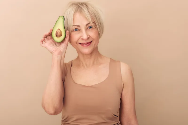 Smiling mature woman with short blonde hair holds half of fresh avocado near face on beige background. Concept of healthy lifestyle, natural nutrition, wellness, and skin care for seniors.