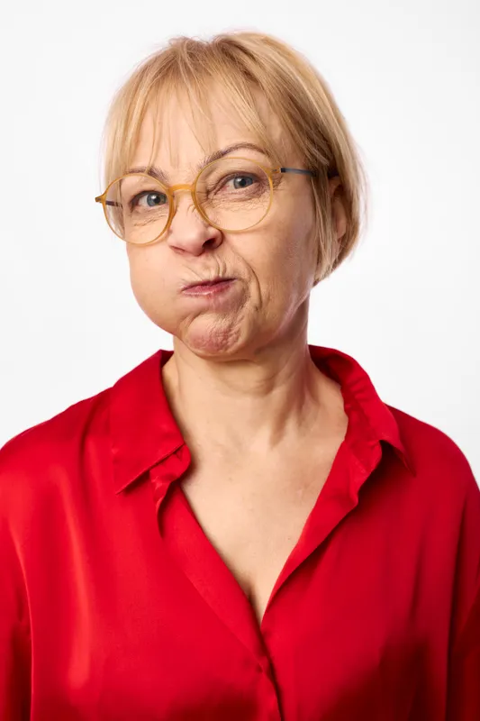 Senior woman portrait with glasses and blonde short hair, wearing red shirt, close up face in white studio, calm neutral expression, minimal background