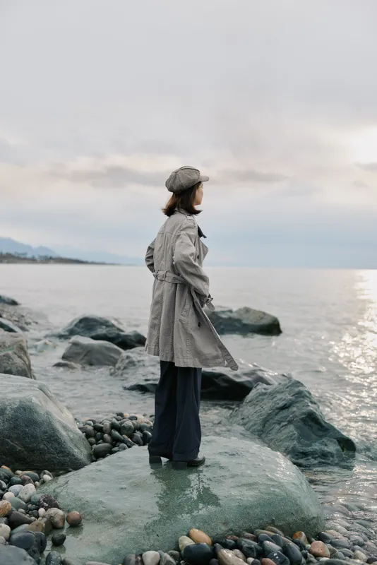 A woman in a checkered coat and hat stands on a rocky shore looking at the calm sea under cloudy sky. The scene depicts solitude, nature, and peaceful contemplation.