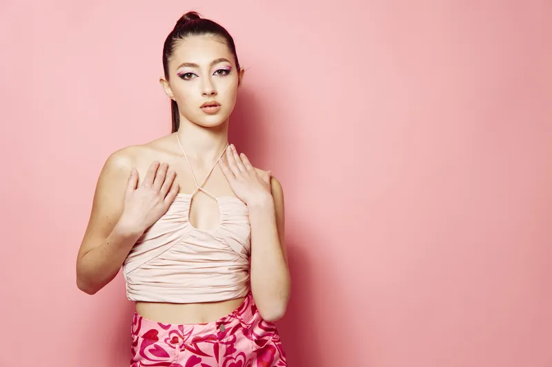 Young woman in trendy fashion posing against pink background with neatly tied hair makeup struck by soft lighting. Concept for beauty creative and fashion.