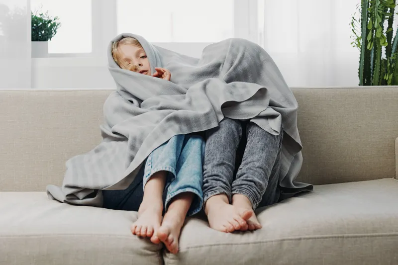 Two children sit barefoot on a beige sofa wrapped together in a grey blanket. Natural light from window and green plants create a cozy home atmosphere of comfort and warmth.