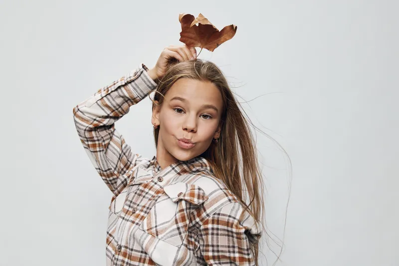 Girl holding dry autumn leaf above head and posing on white background. Child wearing plaid shirt with long hair making playful expression and having fun with nature element indoors.