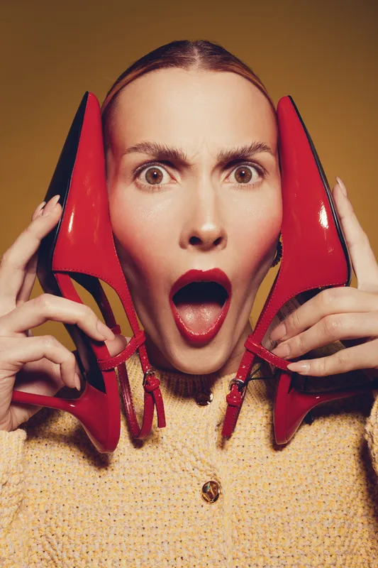 Surprised woman holds red high heel shoes near face with open mouth. Close-up portrait on yellow background. Concept of fashion, style, footwear, and female expression.