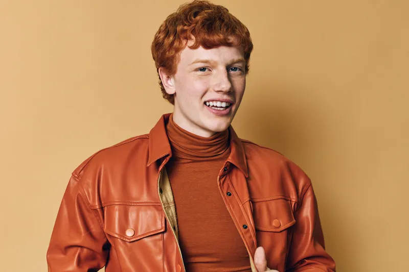 Young man with red hair wearing brown leather jacket and turtleneck smiling with confident expression against beige background in studio portrait.