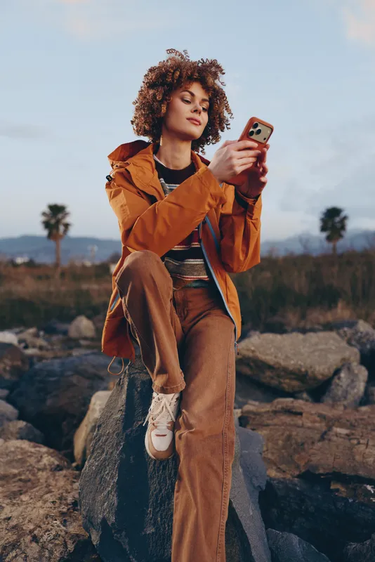 Woman lifestyle outdoors selfie phone coat, curly hair smile, standing on rocky ground at sunset, relaxed walking pose, natural light, cinematic colors, travel mood, fresh breeze