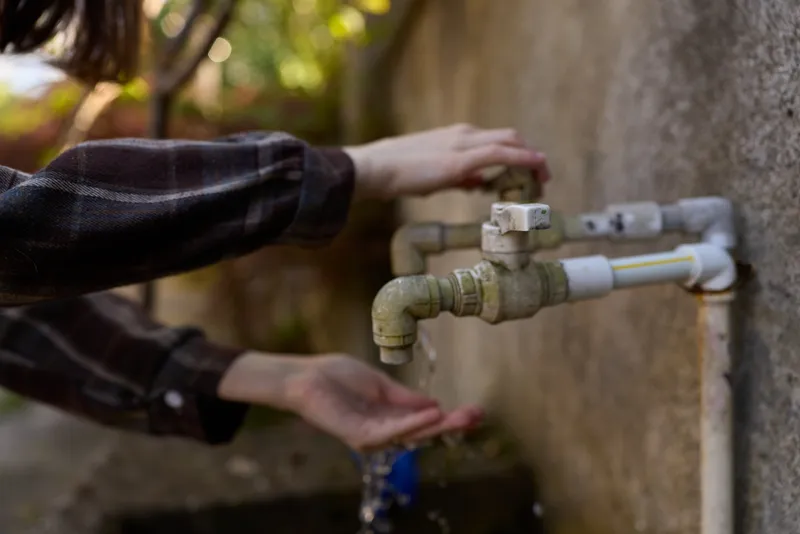Woman lifestyle scene shows hands at an outdoor faucet beside a wall, washing under running water, a simple casual moment captured in a practical outdoor setting with natural light