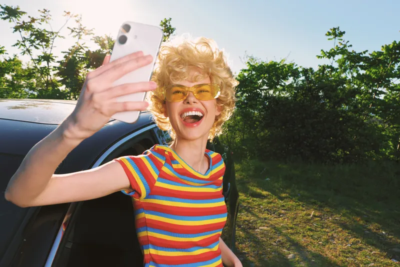 Young woman with blond hair in a striped shirt takes a selfie beside a car, radiating a playful retro vibe and a carefree summer mood.