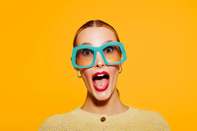 Surprised woman with blue oversized glasses and red lipstick looks excited on bright yellow background. Concept for fun, fashion, emotions, and colorful style portrait.