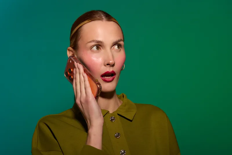 Surprised woman with light skin and blonde hair holds smartphone to ear, wearing green shirt. Facial expression shows shock or disbelief on solid green background. Concept of communication and