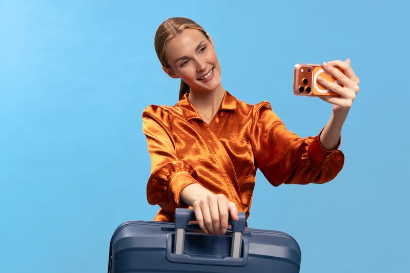 Young woman in orange blouse takes selfie with smartphone while holding suitcase handle. Blue background studio shot. Concept for travel, vacation, and social media lifestyle moments.