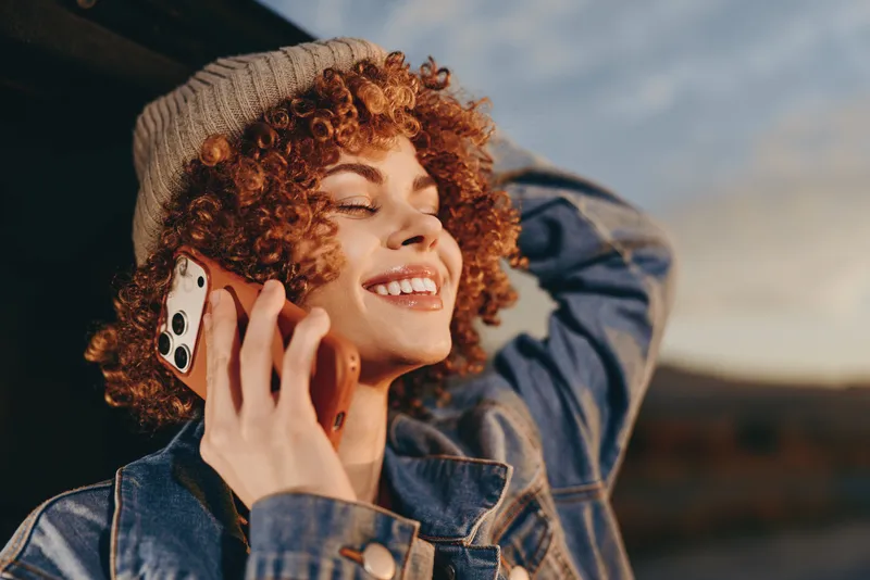 Woman with curly red hair smiles while making a phone call outdoors, wearing a denim jacket, casual lifestyle portrait in bright natural light, relaxed carefree mood today.