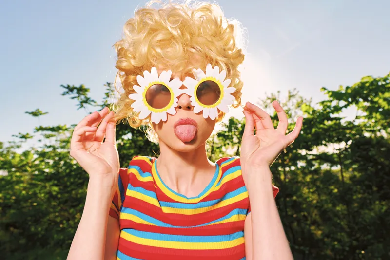 Cheerful woman outdoors, wearing flower sunglasses and striped top, enjoying a sunlit moment with playful pose, retro film color and natural tones, lush greenery background, carefree mood