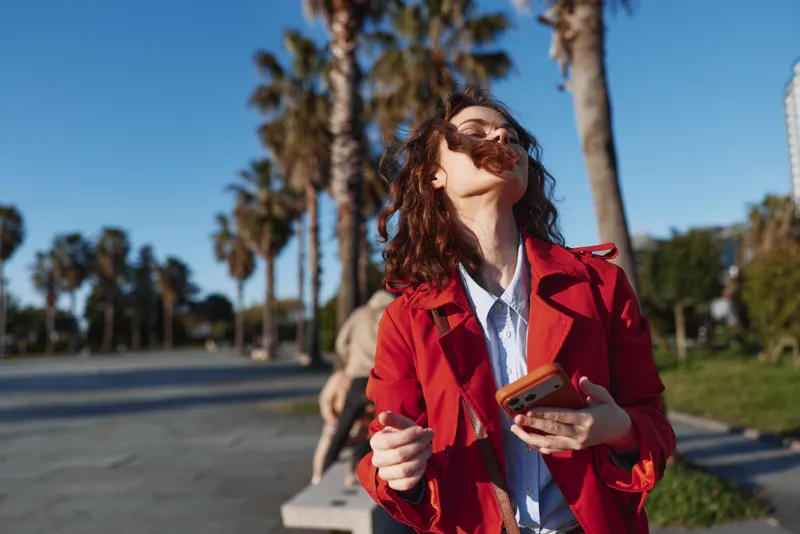 Young woman outdoors in red coat, enjoying sunny day, holding smartphone, hair blowing in wind, palm trees background, casual urban setting