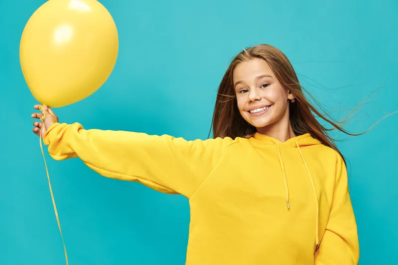 Smiling girl wearing yellow sweater holding yellow balloon on blue background with hair blowing and cheerful expression for happiness and celebration concept.