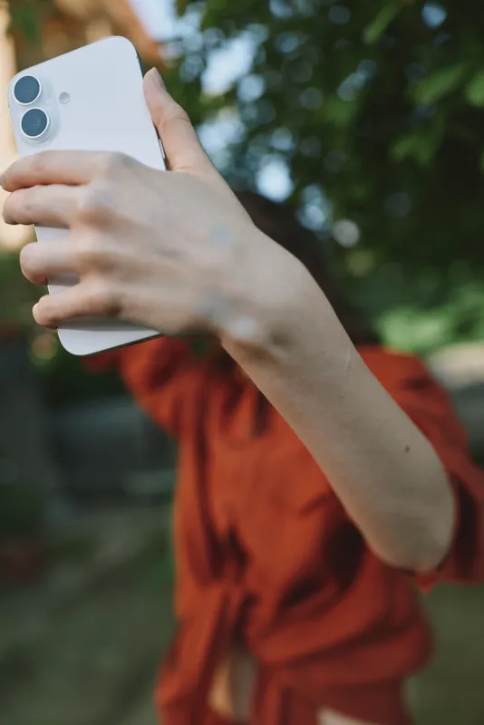 Casual woman outdoors, holding a smartphone for a selfie, sunlight and greenery in soft colors, relaxed pose, park background, natural lifestyle moment