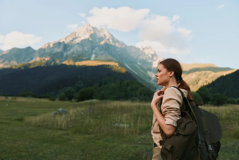 Young woman traveler with backpack standing in green meadow looking at mountain landscape, clear sky, outdoor adventure, nature exploration, calm moment