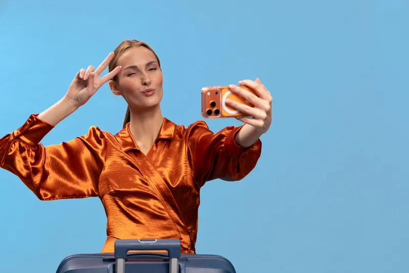 Young woman in orange shiny blouse takes selfie with smartphone showing peace sign. Blue background and suitcase visible. Concept of travel, vacation, and lifestyle.