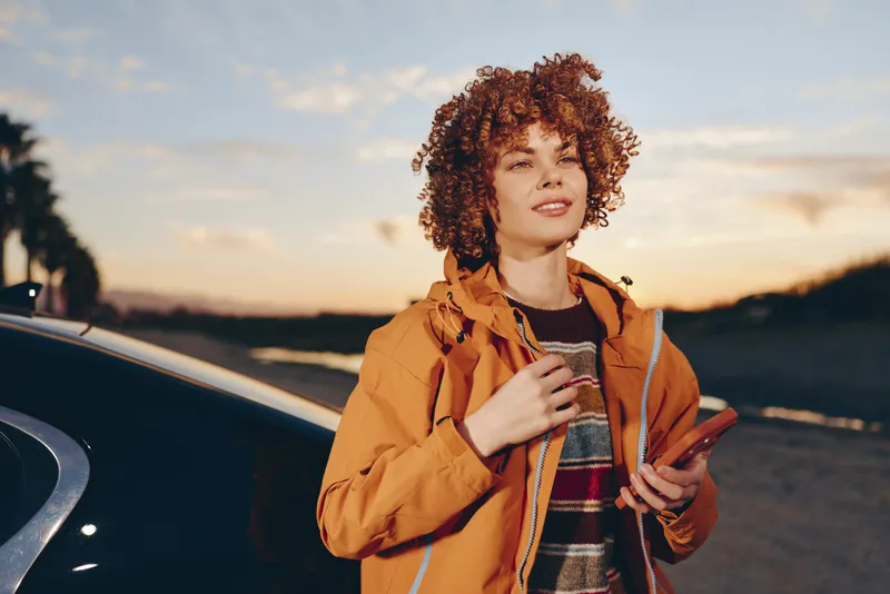Woman lifestyle sunset car travel road jacket outdoors curly hair, standing by a car on an open road, orange jacket, holding a bag, smiling softly, peaceful warm evening light outdoors