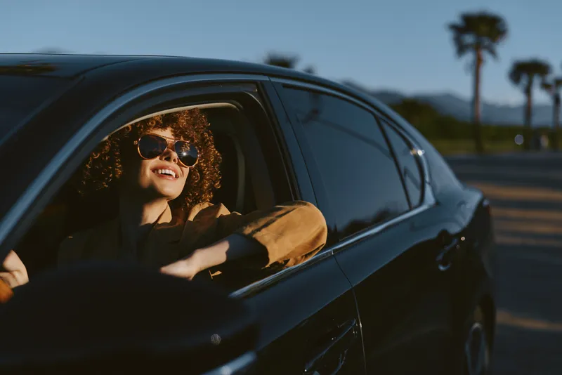 A smiling woman leans from a sleek car window in warm light, sunglasses on, hair catching the breeze. Concept: casual travel, modern lifestyle, carefree road outing.