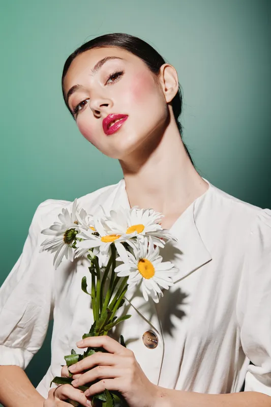 Young woman with shiny bold red lipstick and natural glowing skin holds white daisies. Styled with sleek hair and an elegant white outfit on muted green background. Beauty fashion concept.