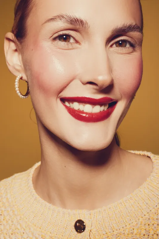 Close-up portrait of a smiling woman with red lipstick and hoop earrings wearing a knitted sweater. Warm lighting highlights her clear skin and joyful expression on a yellow background.