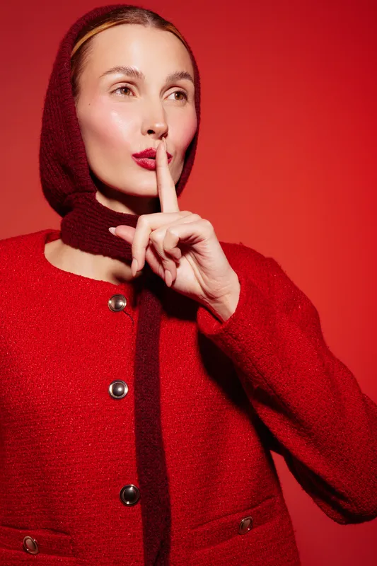 Woman in red woolen coat and matching headscarf holds finger to lips in shushing gesture against red background. Concept of silence, secrecy, and quiet communication.
