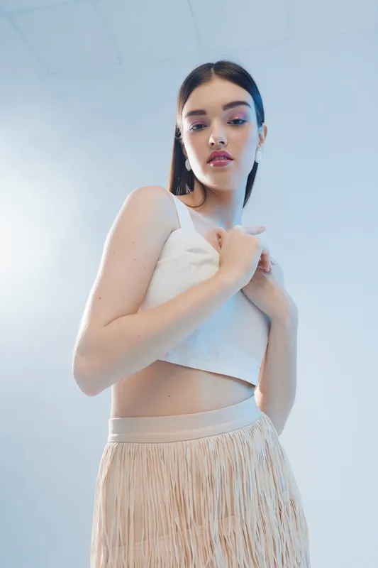 Young woman with long dark hair wearing a white crop top and beige fringe skirt poses with hands near chest. Studio shot with soft lighting on light blue background. Concept of woman beauty fashion.