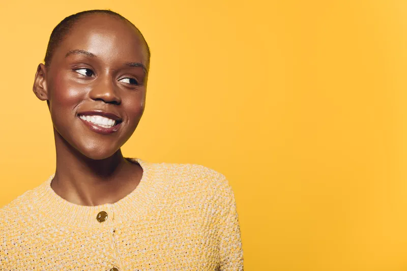 Smiling young black woman with dark skin wearing yellow sweater looks to the side. Warm studio lighting and solid yellow background create a cheerful and positive atmosphere.