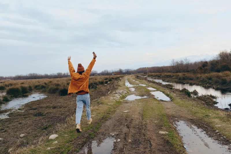 Woman lifestyle outdoors walking on muddy path in countryside nature, joyful moment, arms up, long hair in coat, spring morning, wide horizon, empty trail near river, cloudy sky