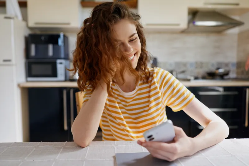 Woman lifestyle at home, smiling while using a smartphone in a bright kitchen, striped tee, relaxed mood, everyday moment, indoor setting, cheerful vibe, candid moment.