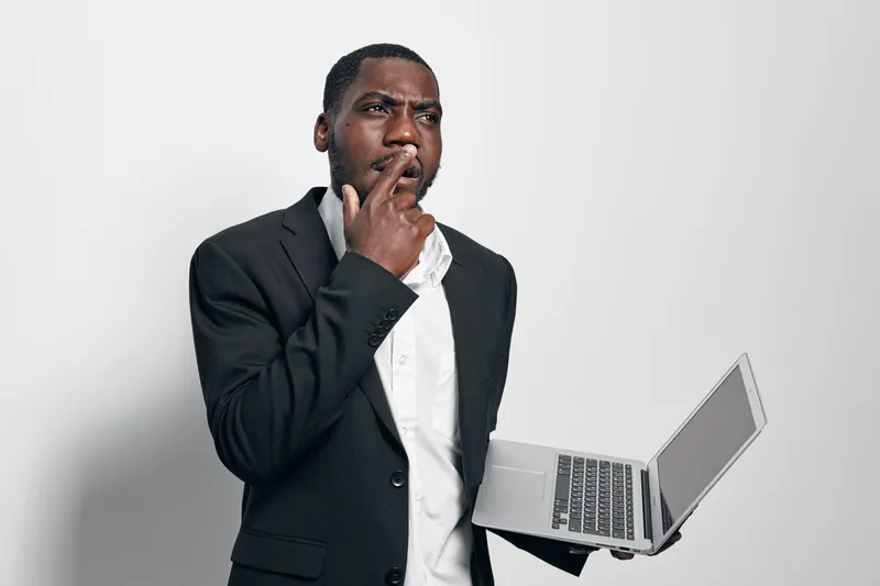 African American man with dark skin in black suit holding laptop and thinking with finger on lips isolated on white background for business or technology concept.