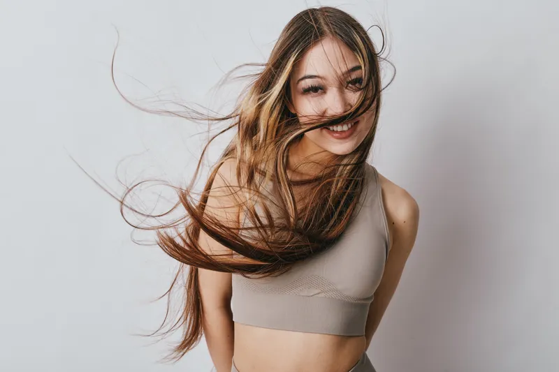 Woman smiling with long hair flowing in motion wearing sleeveless top standing against plain background, youthful energy and happiness captured in studio portrait session.