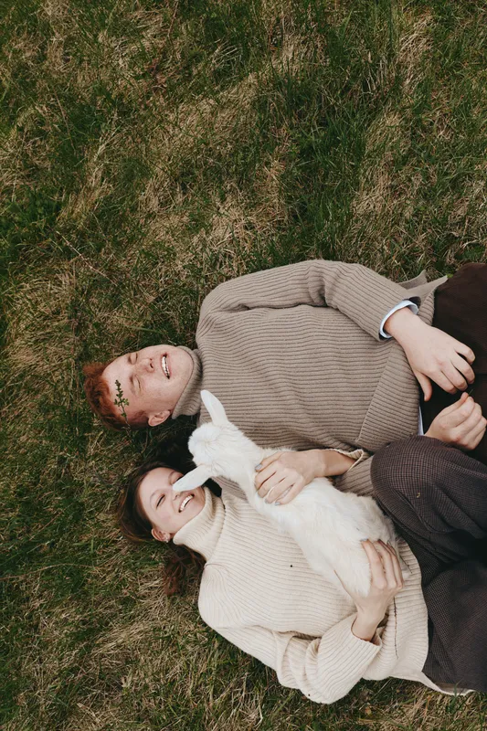A goat and kid lie beside a relaxed couple in an outdoor grass scene, capturing lifestyle, affection, and companionship in a calm, natural family moment outdoors.