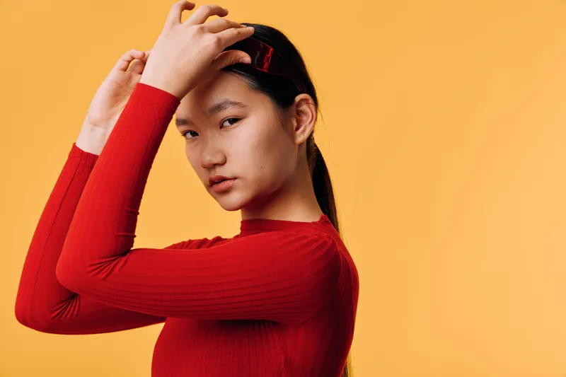 Young woman wearing red sweater adjusting hair clip with serious expression on yellow background. Asian female model looking at camera in casual style portrait with minimal makeup and accessories.