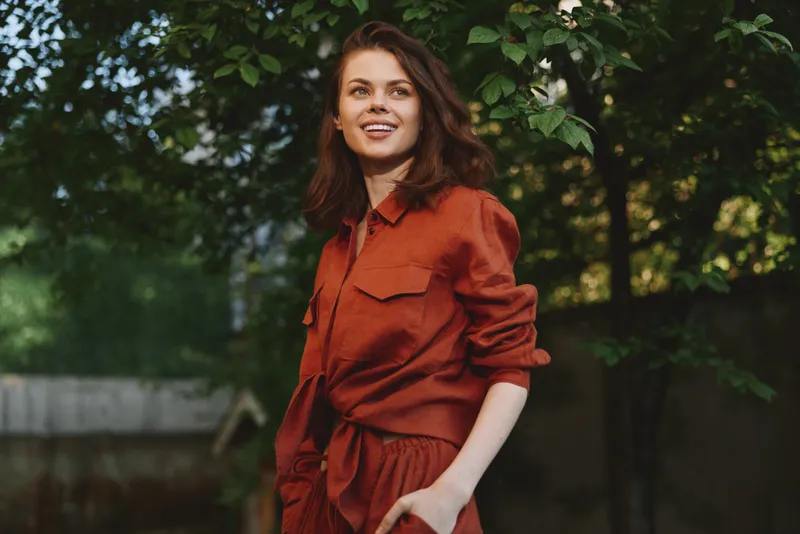 Young woman in a rust jumpsuit smiles outdoors among green trees, presenting a casual lifestyle moment with relaxed confidence and natural charm