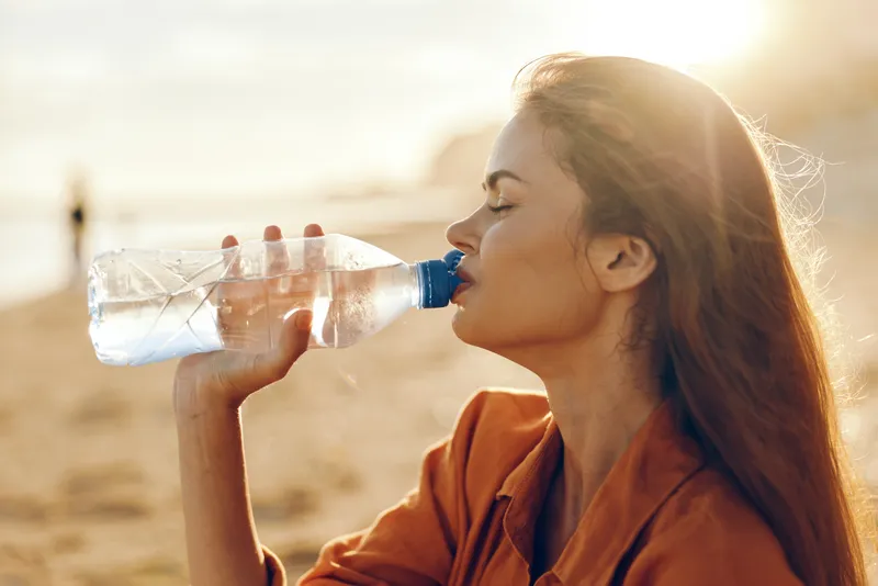 Woman drinking water from bottle outdoors in sunlight, hydrated for health, calm smile, natural warm tones, sandy background, summer breeze, after workout