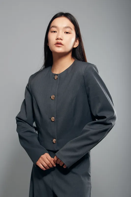Young woman with long dark hair wearing a gray buttoned jacket stands confidently against a gray studio background. Fashion portrait with natural makeup and calm expression.