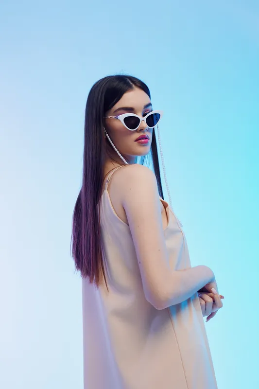 Fashionable woman with long dark hair wearing trendy sunglasses and a beige dress poses against a soft blue background. Concept of beauty, fashion, and style.