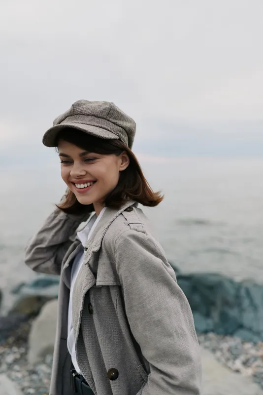 Smiling young woman in grey coat and cap enjoys windy day near rocky sea shore. Casual autumn outfit, natural light, and joyful expression reflect carefree outdoor lifestyle.
