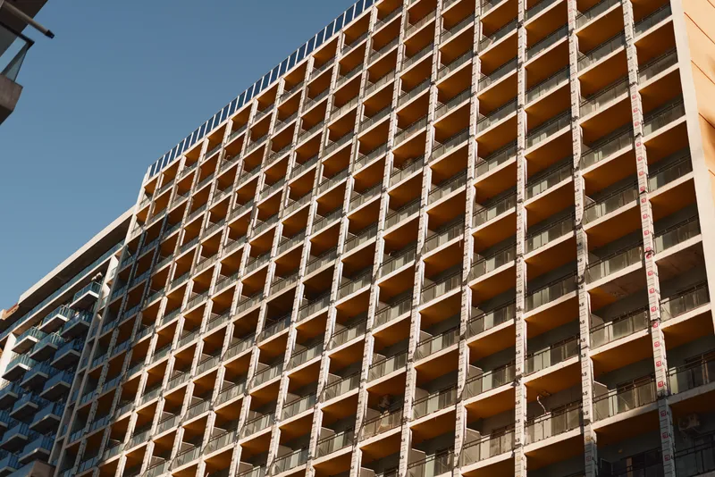 A dramatic upward view of a highrise building with a grid of windows, sunlight casting sharp shadows and emphasizing geometric urban architecture and scale.
