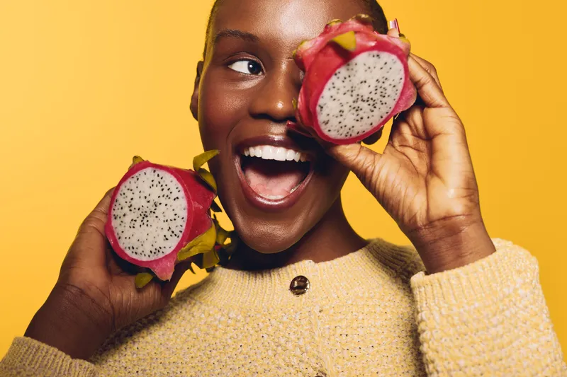 Happy African woman with dark skin holding two halves of dragon fruit near face on yellow background. Concept of exotic fruits, healthy lifestyle, and joyful emotions in studio portrait.