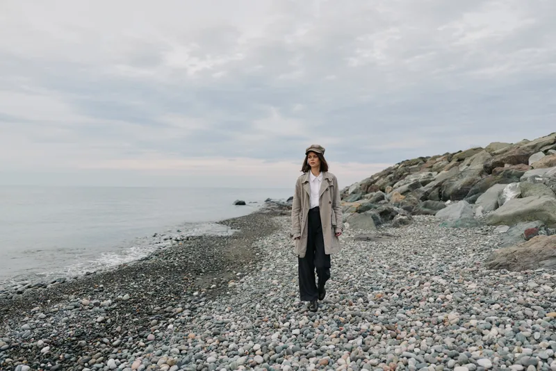 A woman in a trench coat and hat walks alone on a rocky beach under an overcast sky. The calm sea and scattered stones create a peaceful, moody atmosphere of solitude and reflection.