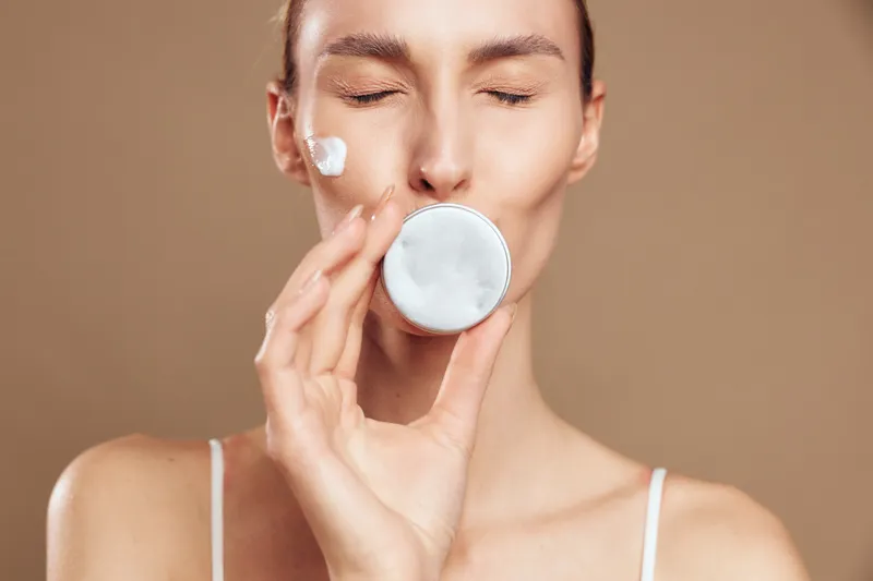 Young woman with closed eyes applying white cream on face, holding a round container, natural skincare routine, soft lighting, neutral background, fresh healthy skin care concept