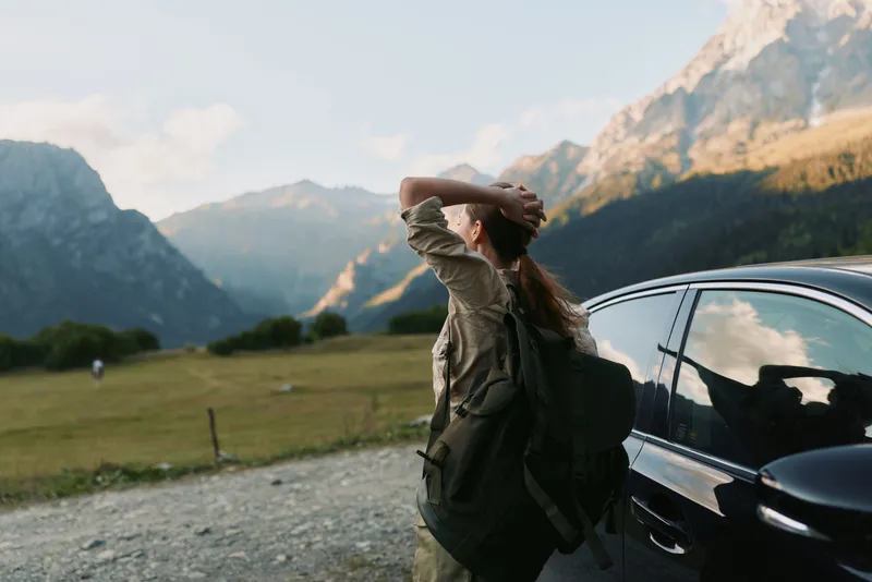 Young woman with backpack stretches her arms beside a parked car in a rugged mountain valley, exuding curiosity and freedom with a sunlit dramatic landscape behind.