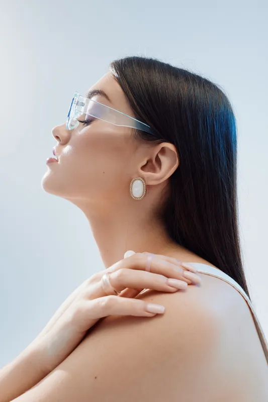 Profile of a beautiful woman wearing transparent glasses and elegant earrings. Her long dark hair is sleek, and she poses with hands on shoulder. Concept of beauty fashion.