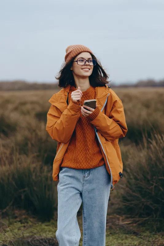 Woman lifestyle portrait outdoors in an autumn field, wearing orange jacket, knit sweater and beanie, smiling and holding a small plant, soft daylight, nature background, calm mood