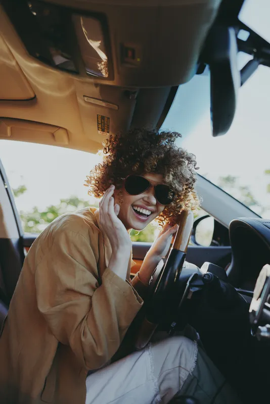 Smiling woman with curly hair wearing sunglasses in a car, capturing a candid lifestyle moment. Warm retro film color tones, natural light, relaxed mood, joyful travel vibe, casual fashion