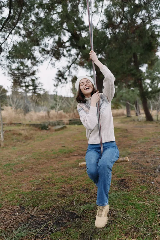 Swinging woman outdoors in a park, smiling and carefree, wearing a sweater and jeans, surrounded by trees and grass, capturing playful recreation and natural daylight.