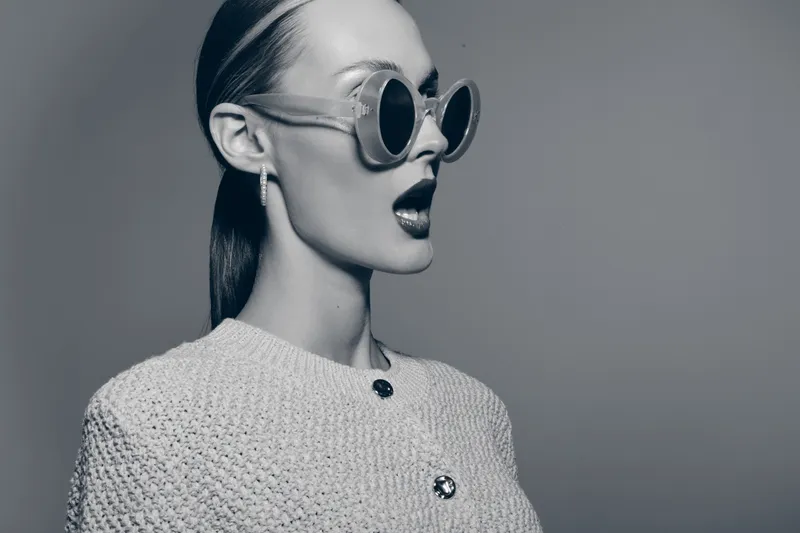 Stylish white woman with pale skin wears large round sunglasses and textured jacket, posing with open mouth. Concept of fashion, style, glamour, and retro aesthetics in studio.